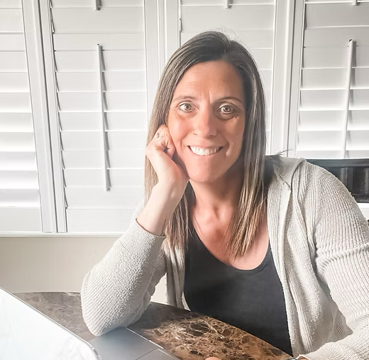 Woman smiling, leaning on her hand, indoors at a table near a laptop and window blinds.
