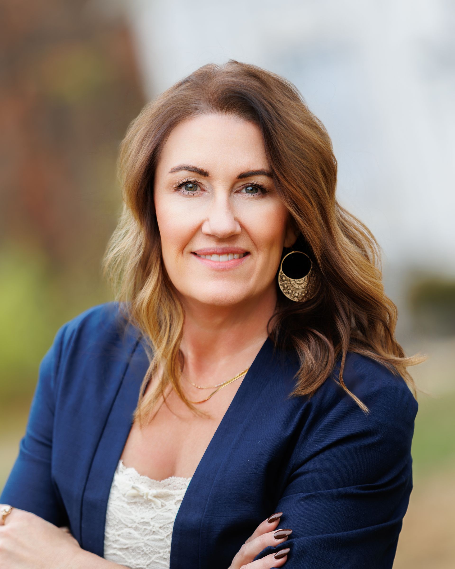 Woman with brown wavy hair smiles wearing a white lace top and jewelry, on a light pink background.