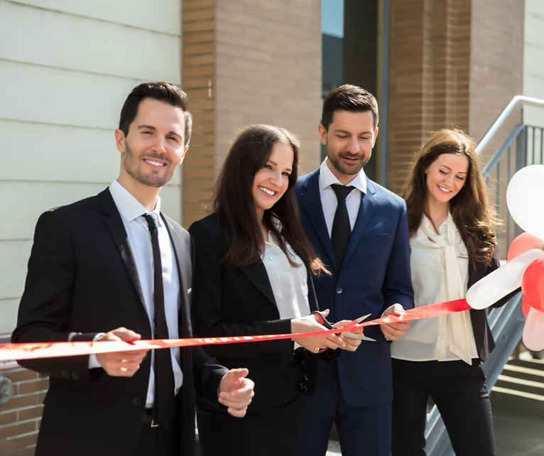 Four people in business attire cutting a red ribbon at a building's opening.