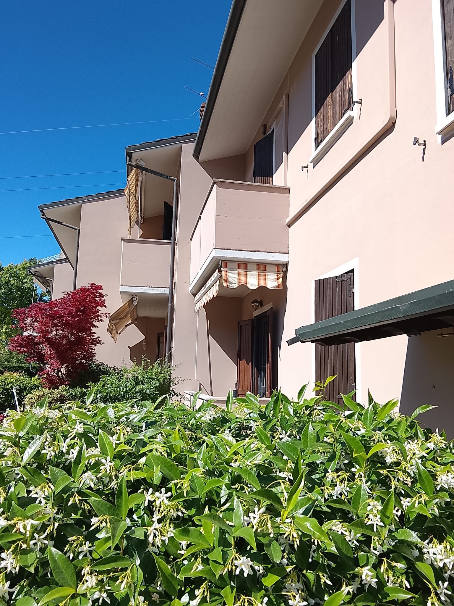 Palazzo di appartamenti rosa illuminato dal sole, con balconi e siepi verdi sotto un cielo azzurro limpido.