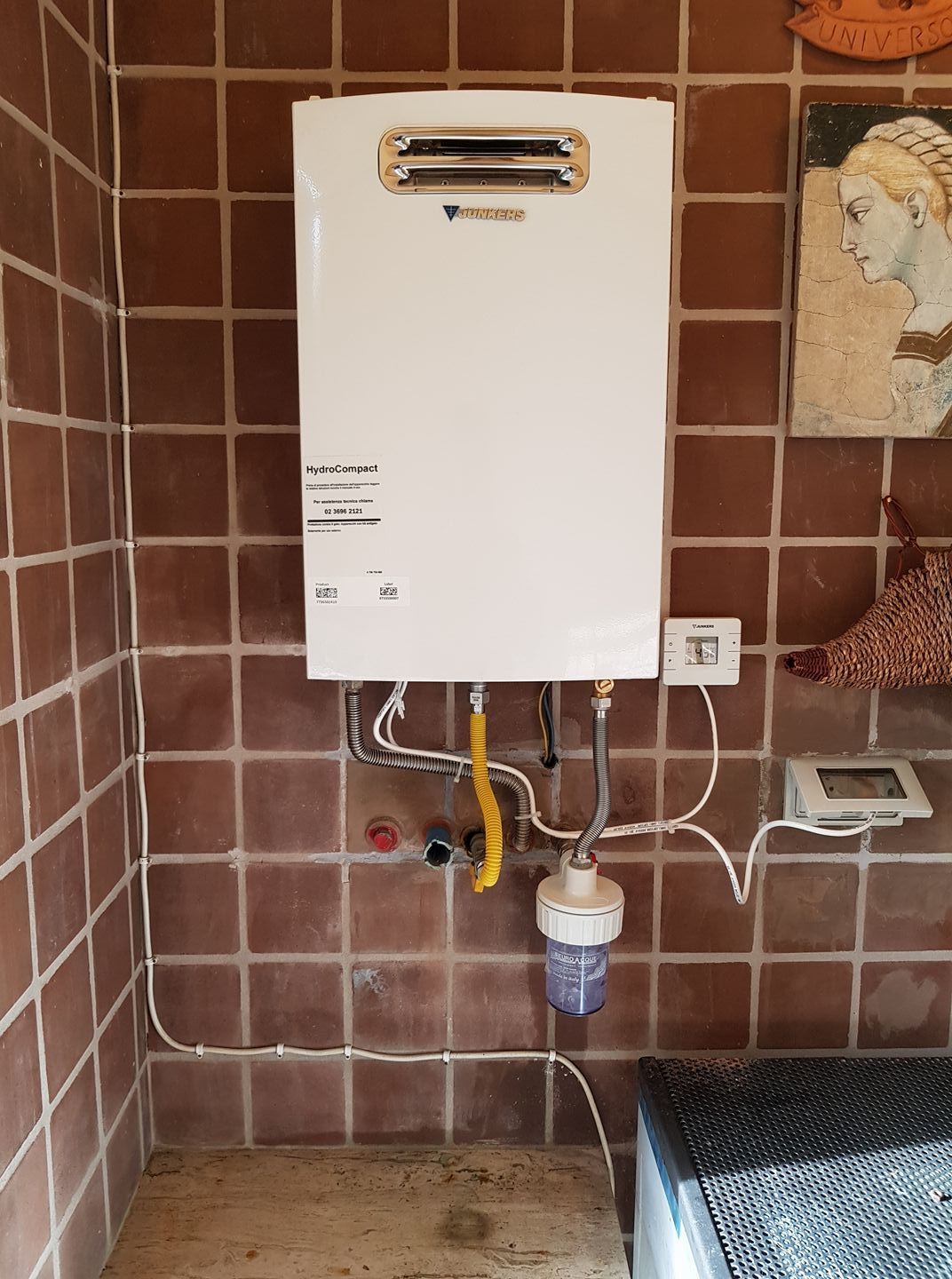 Scaldabagno bianco montato su parete piastrellata marrone con tubo del gas e filtro.