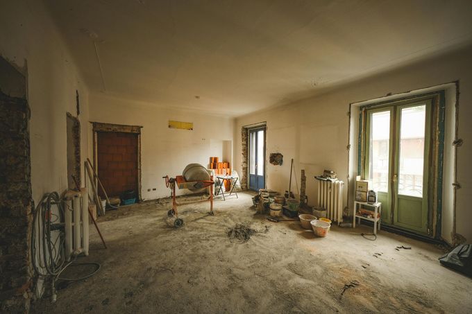 An interior view of a room under renovation with bare walls, dusty floors, a cement mixer, and a set of glass doors.