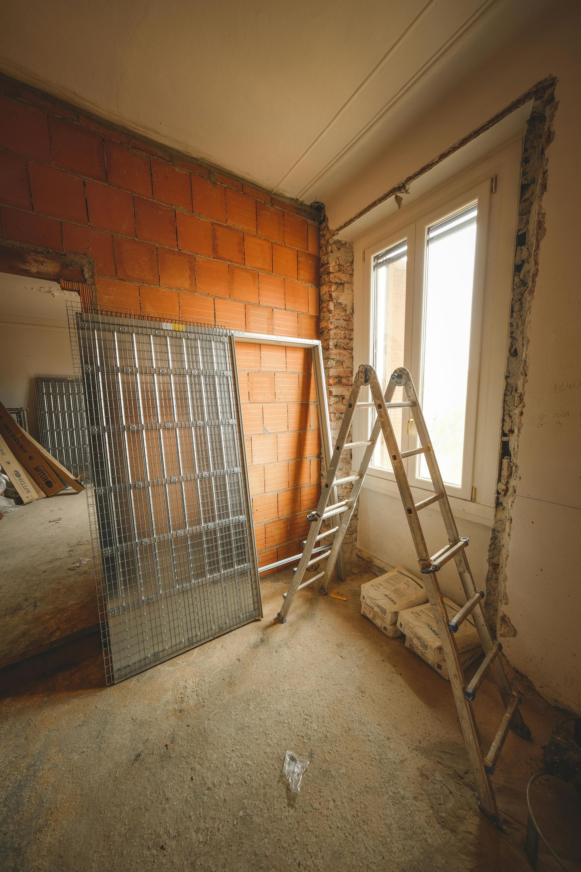 A room under renovation with an exposed brick wall, a metal door frame, and a silver ladder positioned near a window.