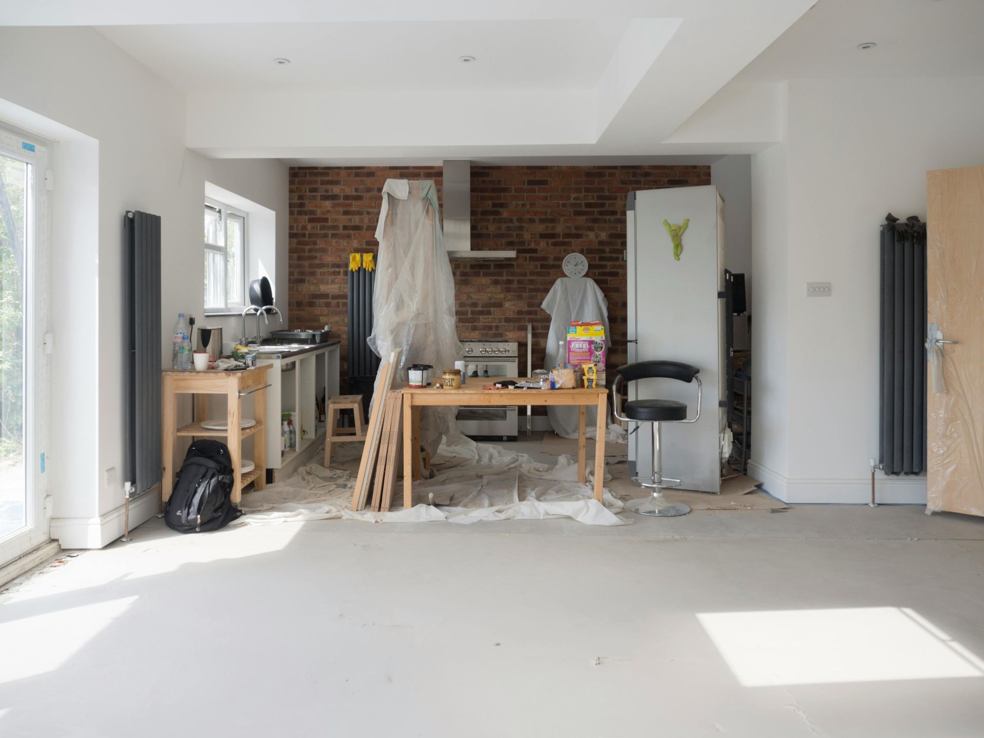 A bright, unfinished kitchen space featuring a brick wall, light wood table, and equipment covered by protective plastic.