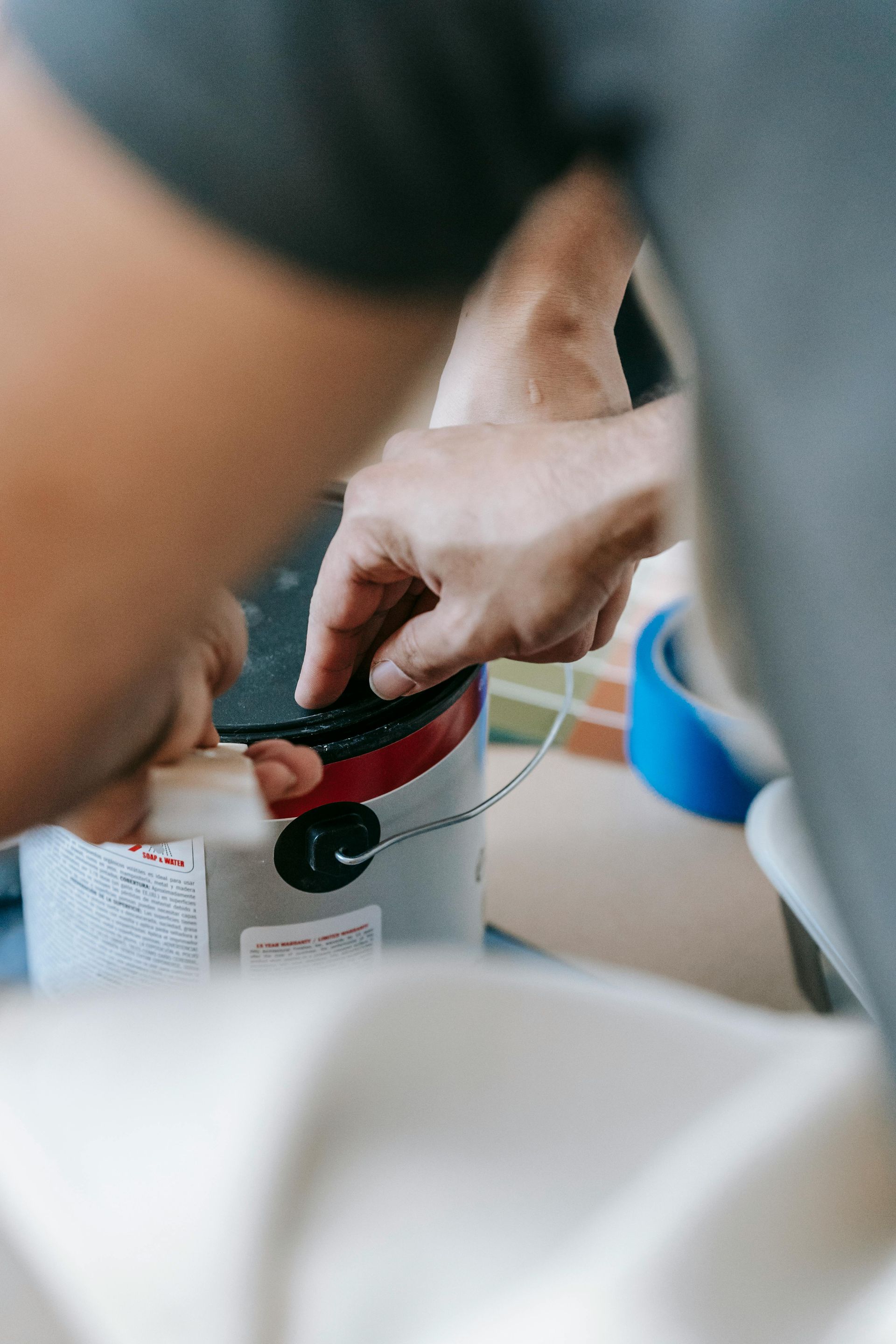 Hands use a tool to open a metal paint can, with blue painter's tape visible on a nearby surface.
