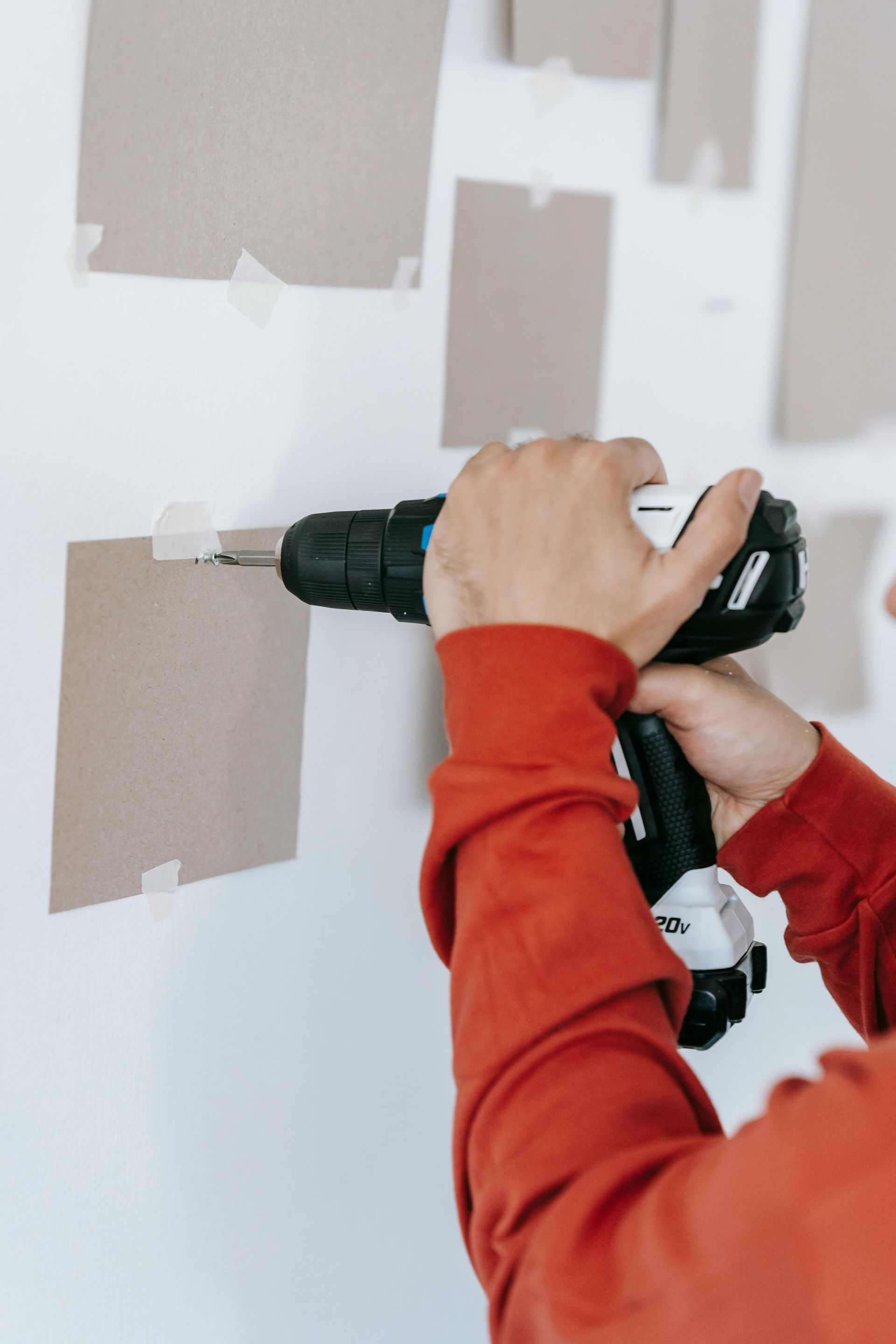 A person wearing an orange long-sleeved shirt uses a power drill to attach a square paper card to a white wall.