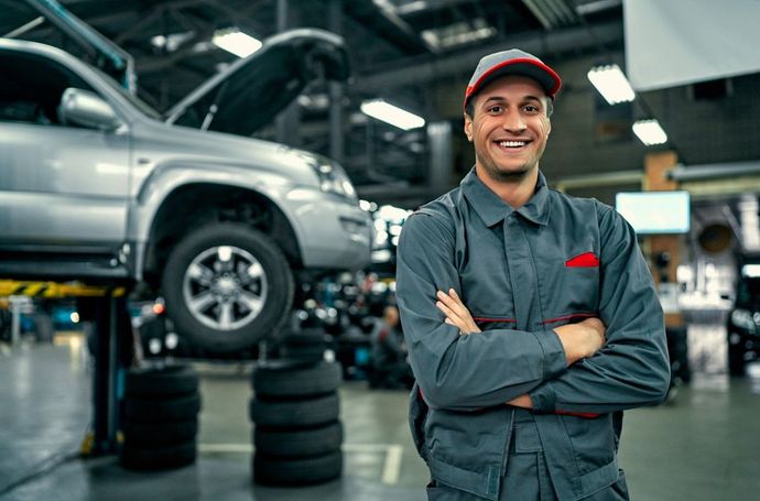 Smiling mechanic in a gray jumpsuit with crossed arms, standing in a car repair shop with a car lifted.