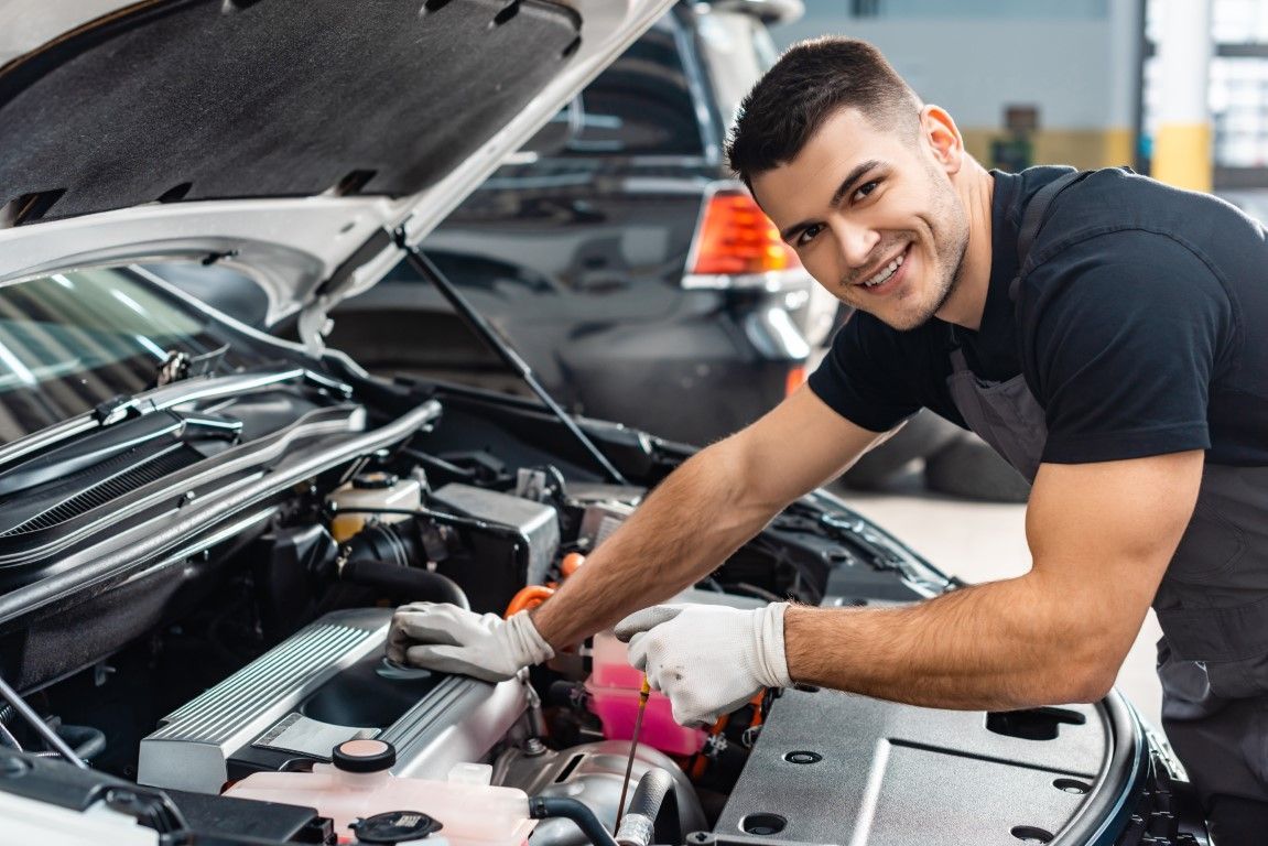 Mechanic in a garage smiling, working on a car engine. He wears gloves, a black shirt, and a grey apron.