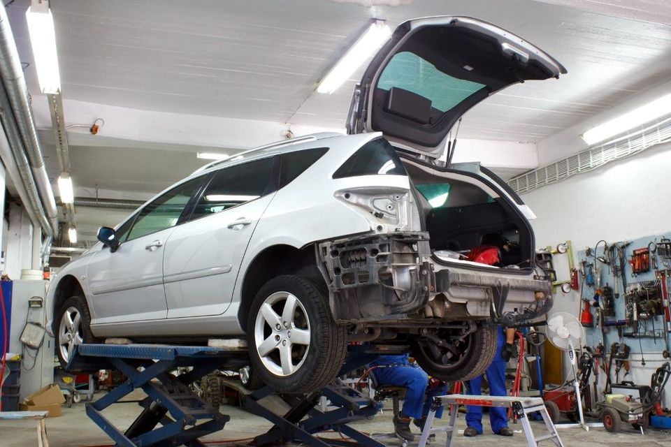 Silver car on a lift in a repair shop, rear bumper removed, trunk open. Tools hang on the wall.