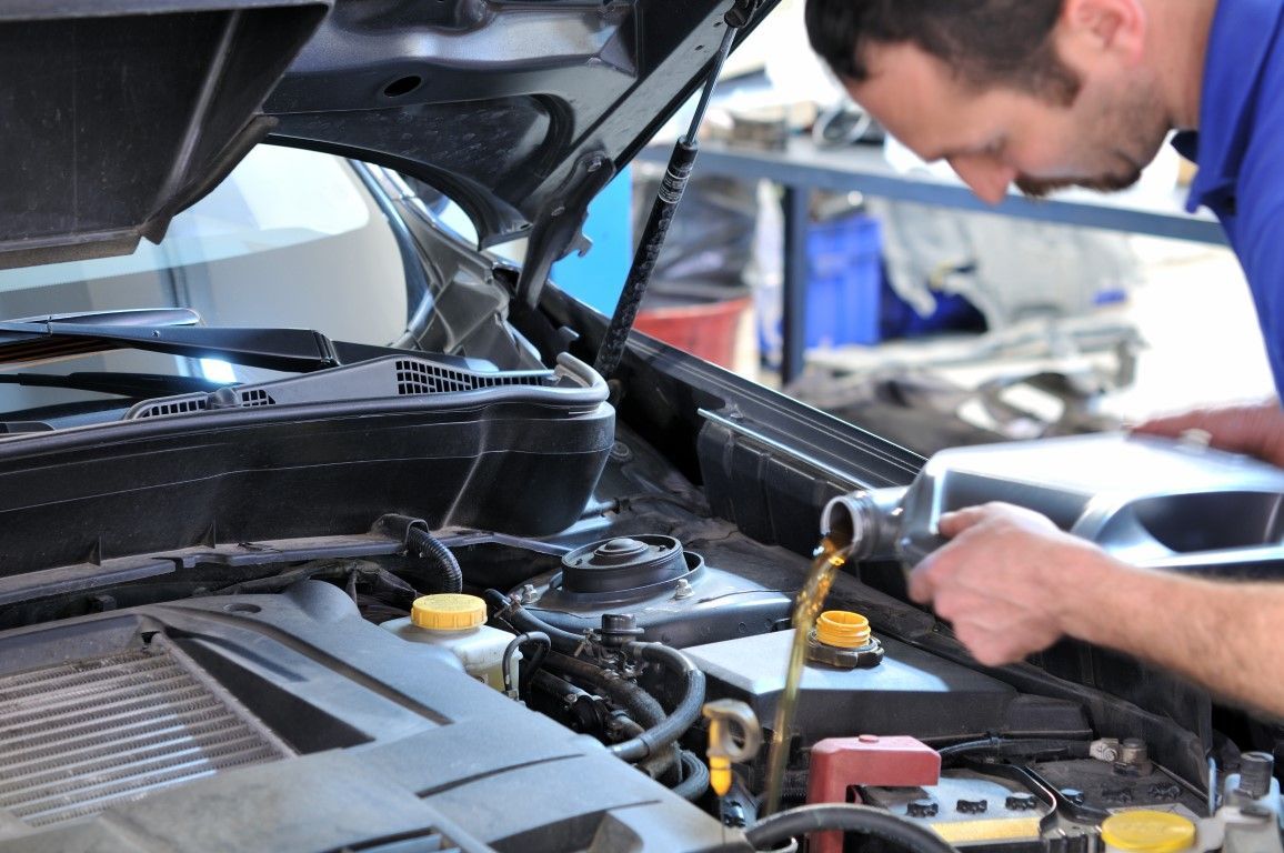 A mechanic pouring oil into a car engine with the hood open. He is wearing a blue shirt.