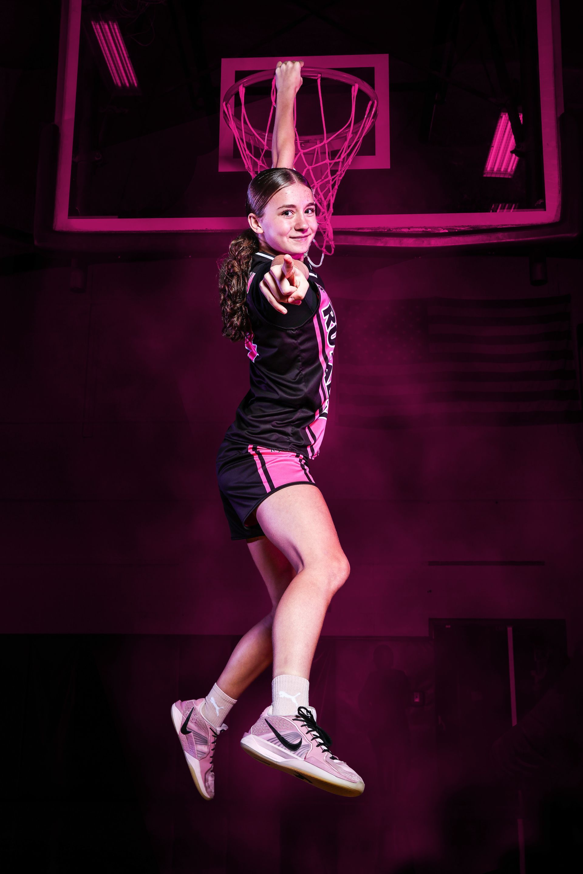 A basketball player jumps to reach a hoop, pointing toward the viewer, bathed in vibrant pink lighting.