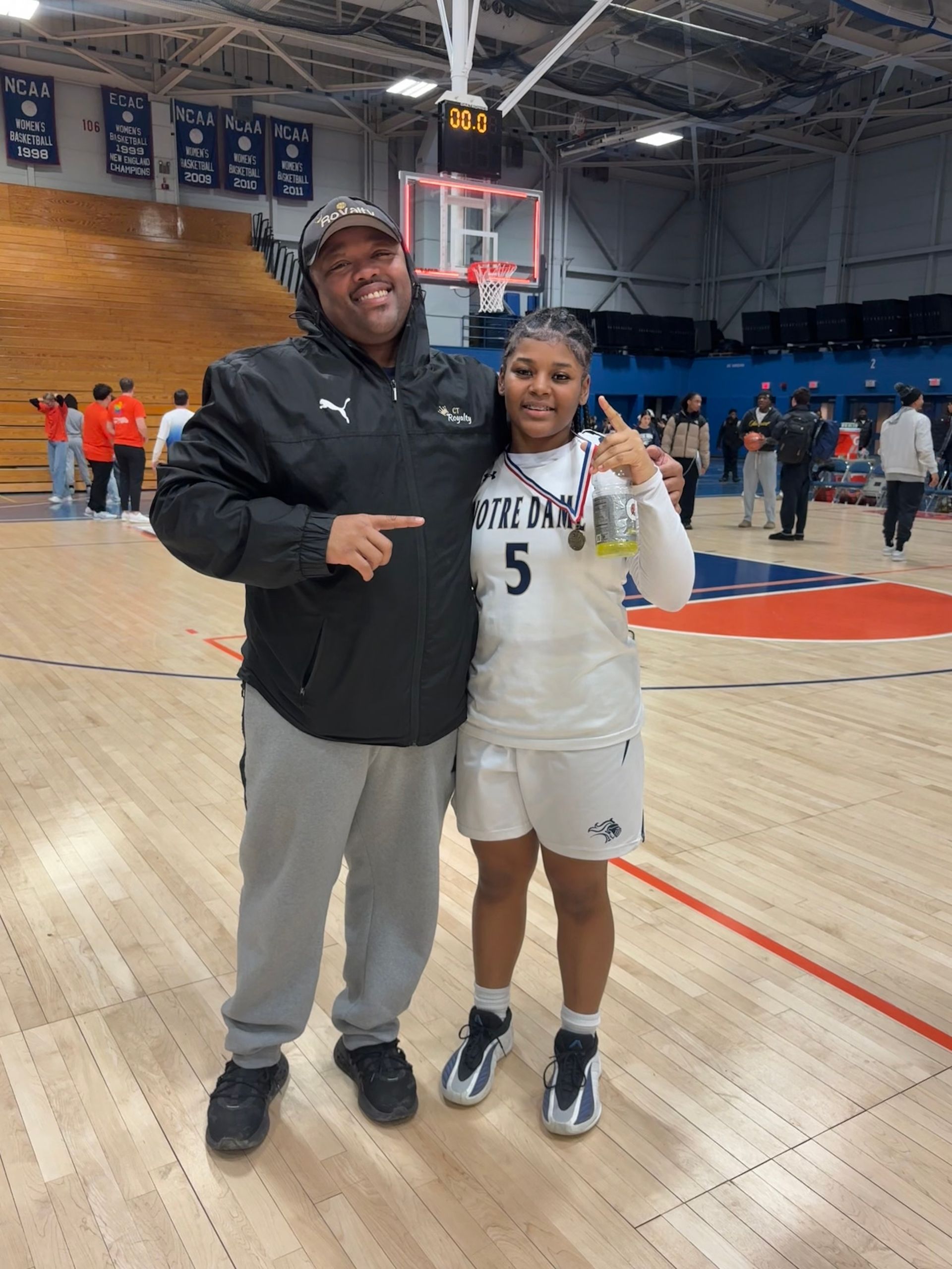A coach in a black Puma jacket stands with a basketball player in a white number 5 jersey on an indoor court.