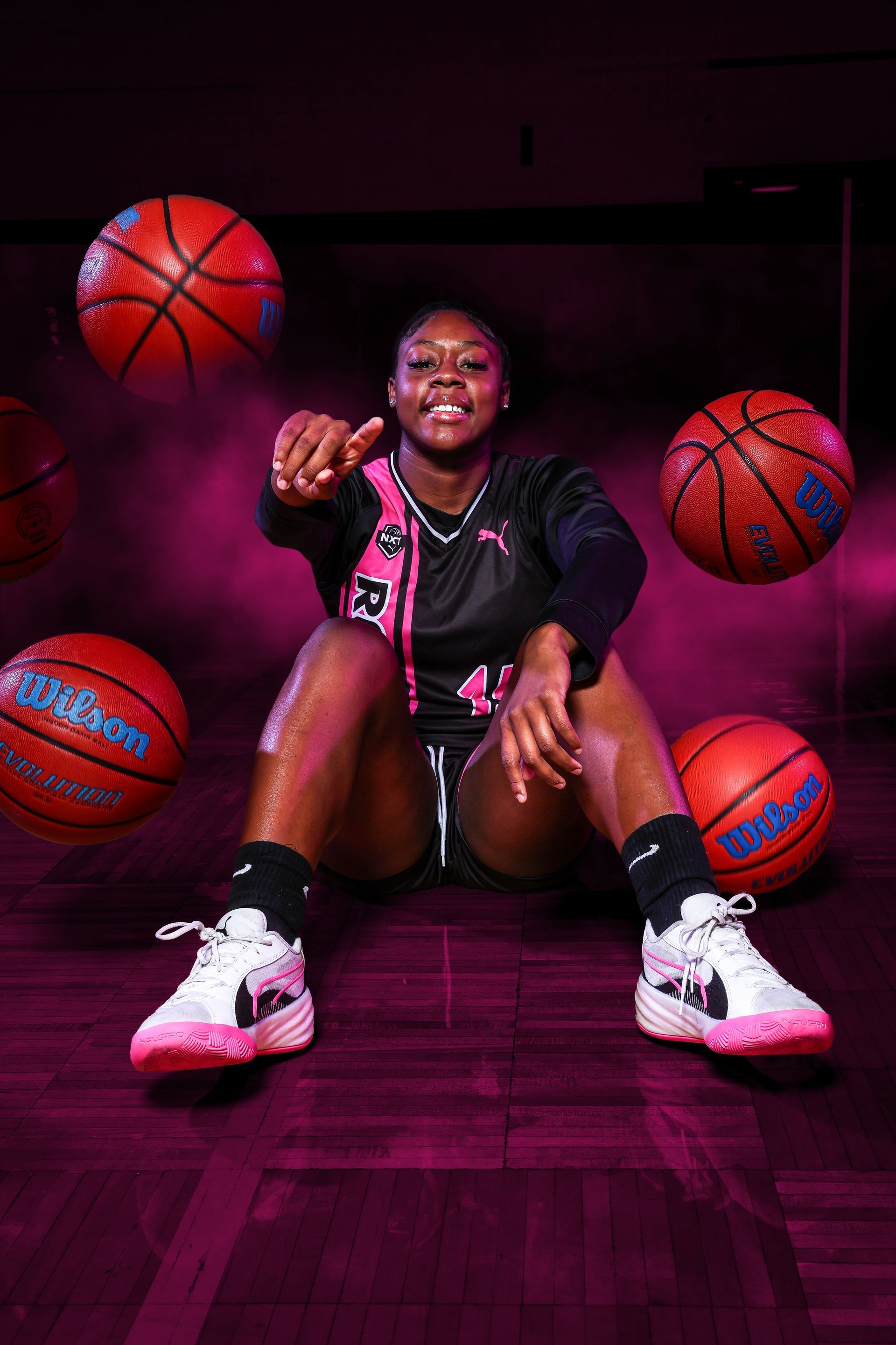 A smiling person in a sports uniform sits on a floor, surrounded by floating basketballs in a dimly lit, purple space.