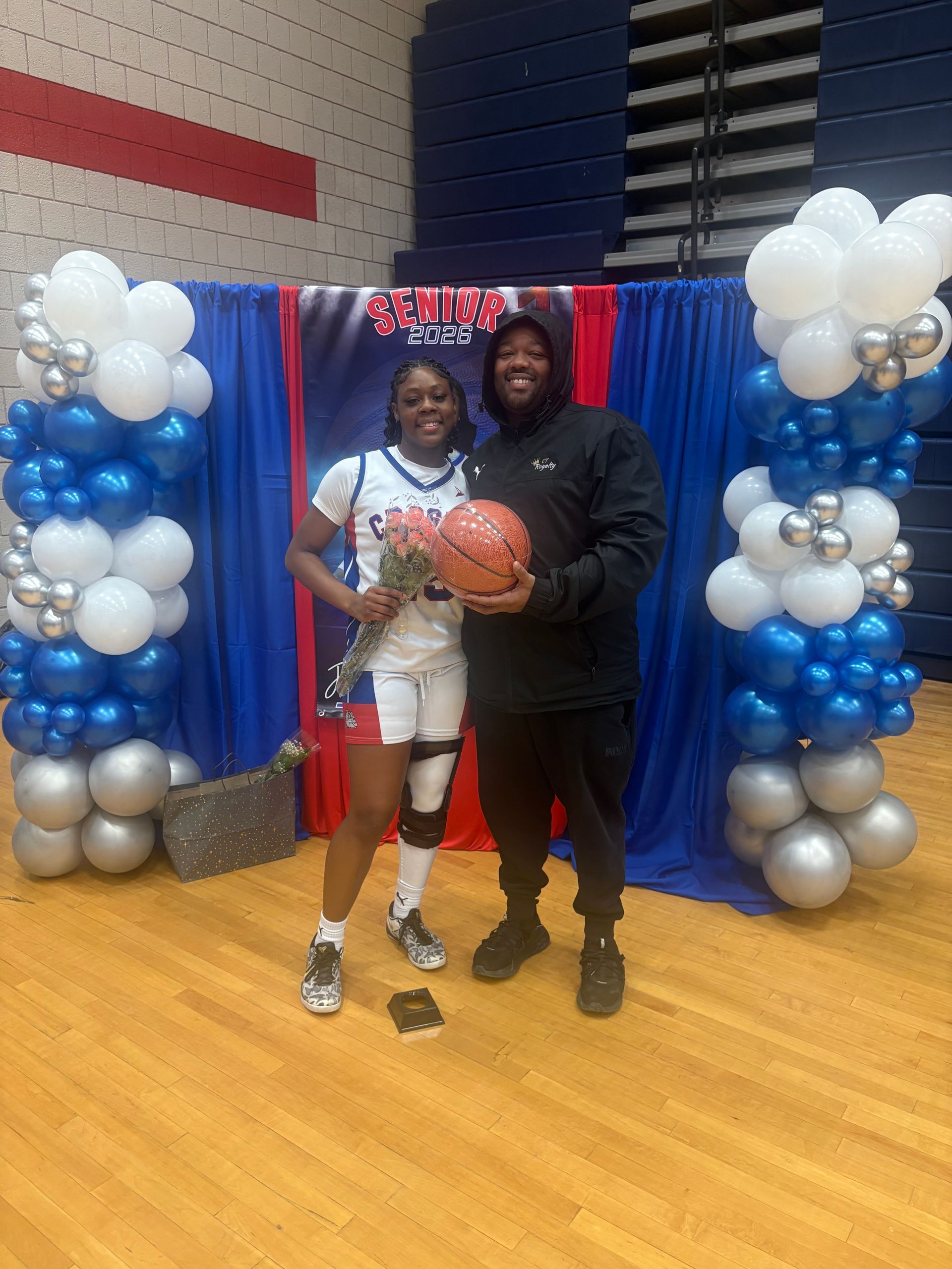 A basketball player holding flowers and a coach holding a basketball stand before a decorated senior night backdrop.