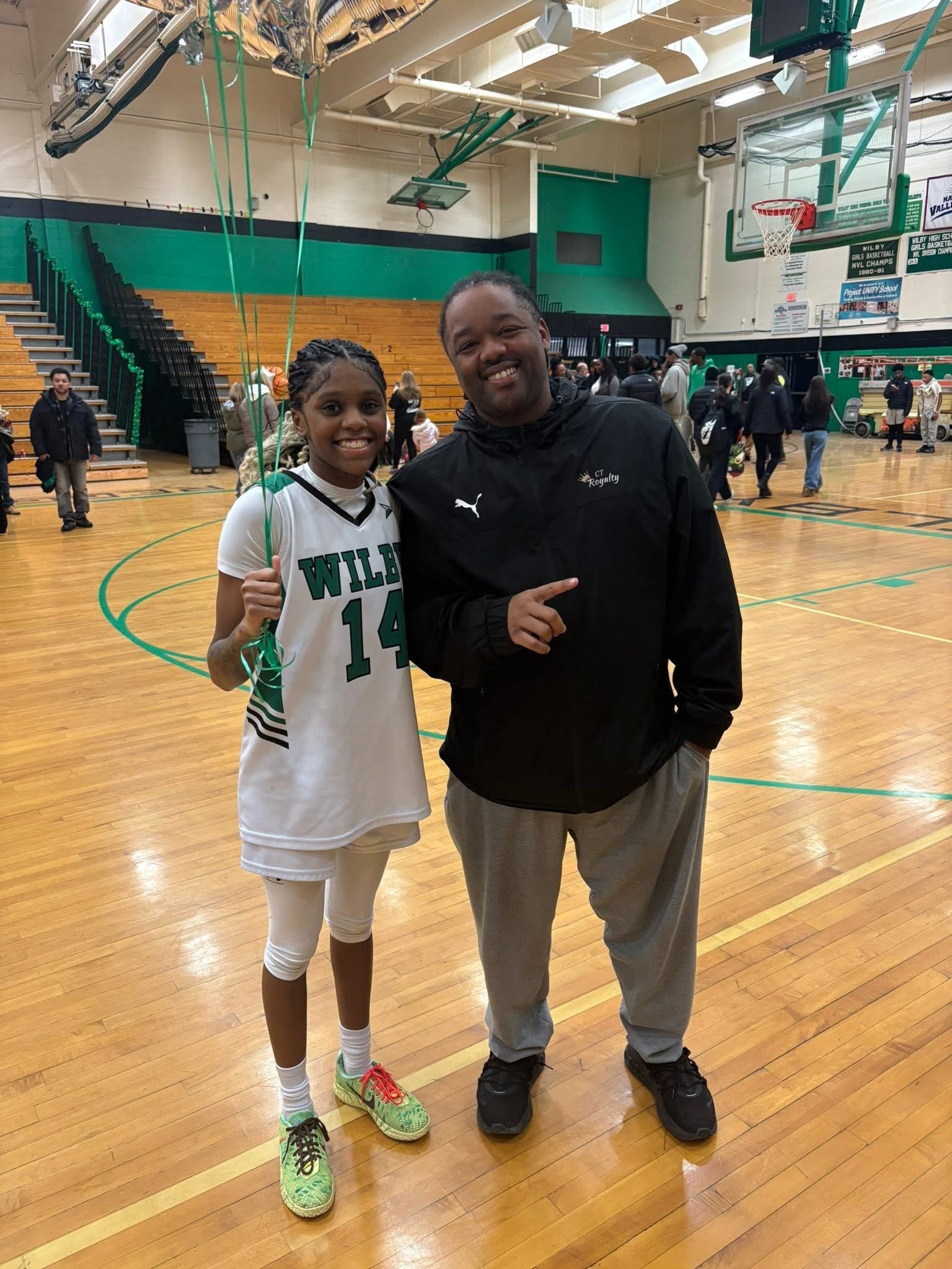 A basketball player wearing a white jersey numbered 14 stands next to a person in a black hoodie on a gymnasium court.