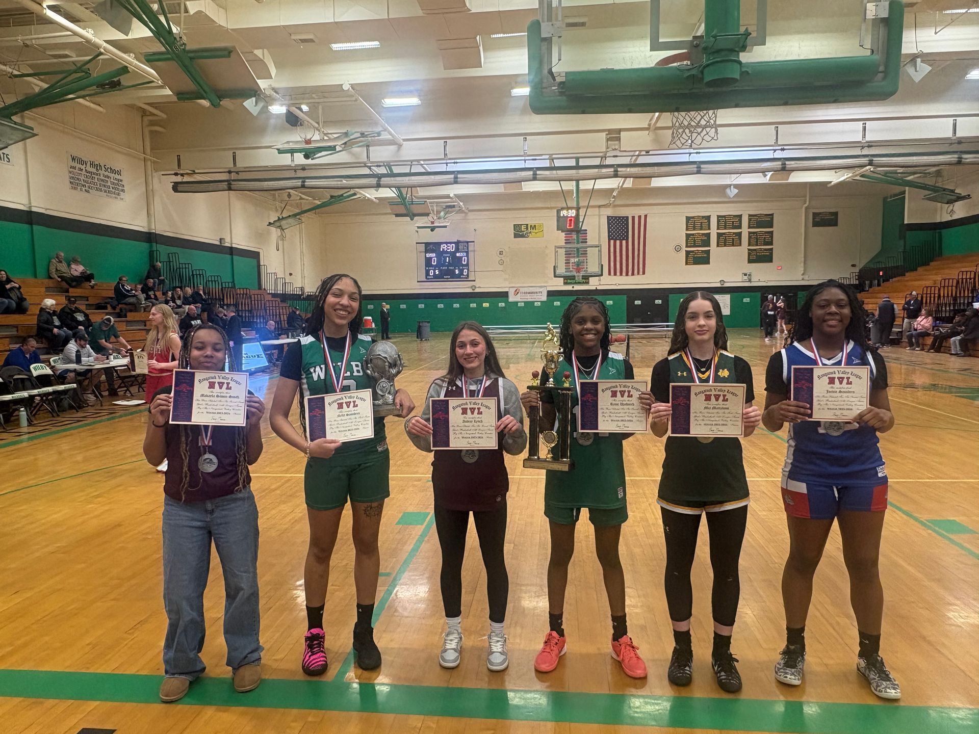 Six basketball players standing on a gym court holding certificates and a trophy after a game.
