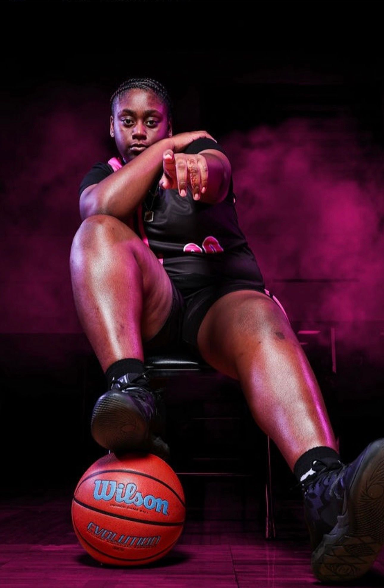 A basketball player in black jersey sits, pointing forward with one foot resting on a basketball against a purple backdrop.