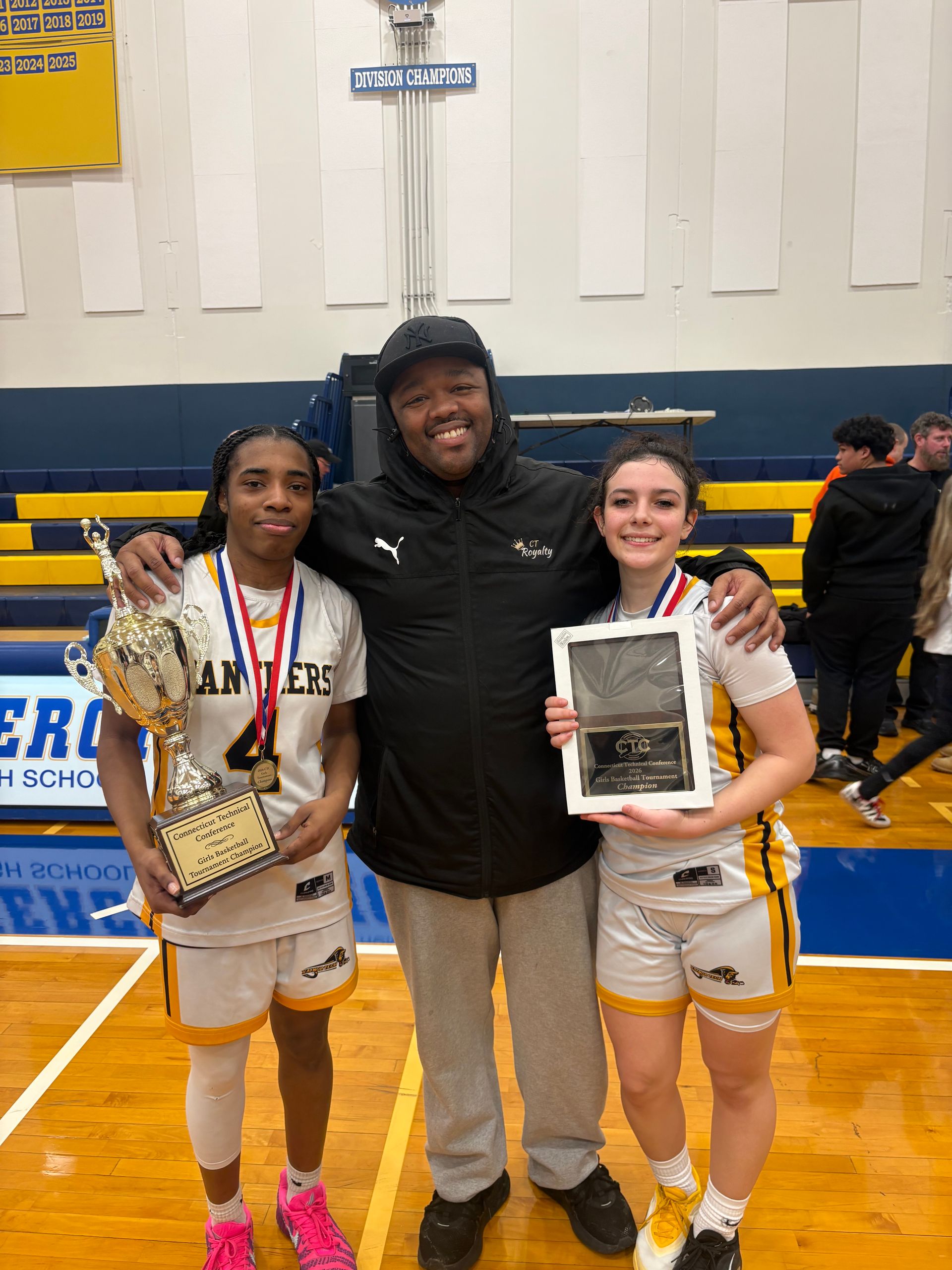 Three people pose smiling in a gym holding a trophy and an award plaque, wearing white and yellow basketball uniforms.