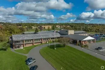Aerial view of a low-rise brick office building with a gray roof and surrounding parking lot under a blue, cloudy sky.