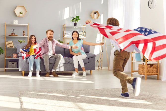 A boy is running with an american flag in front of a family sitting on a couch.