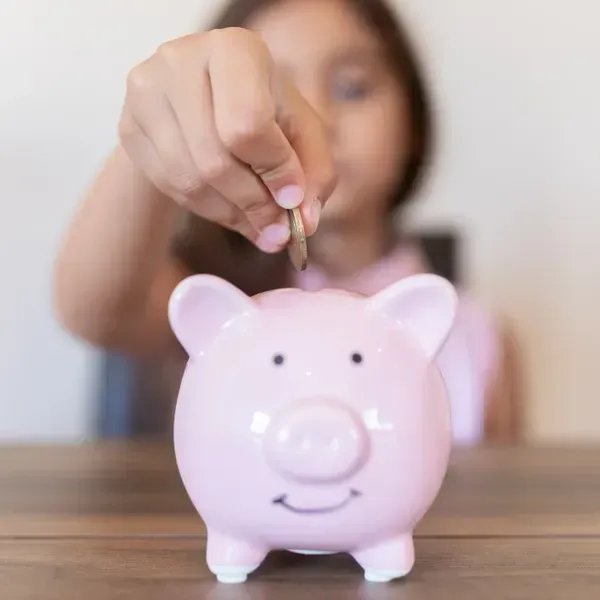 A hand placing a coin into a small, pink piggy bank on a wooden surface.