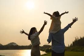 A family with a child on shoulders stands by a lake at sunset with arms outstretched in a joyful, open posture.