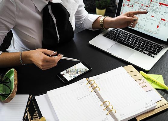 A person pointing at an architectural floor plan on a laptop screen while working at a desk with a notebook and phone.