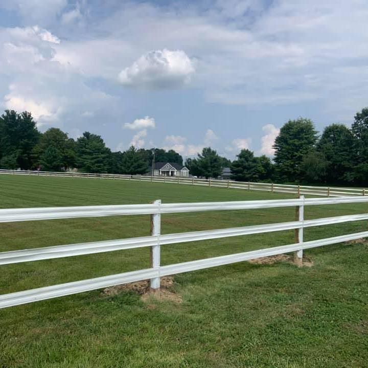 White fence in grassy field, trees and house in background under cloudy sky.