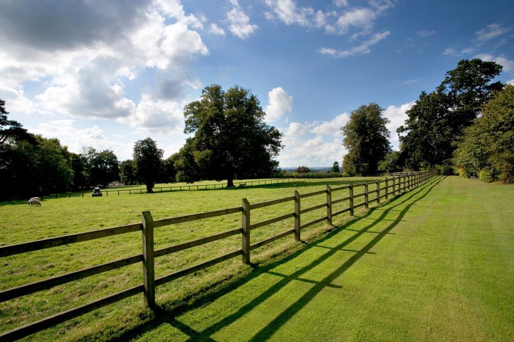 Green field with a wooden fence casting long shadows under a blue, cloudy sky.