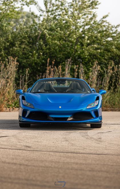 Blue sports car parked on a road, viewed from the front with greenery in the background.