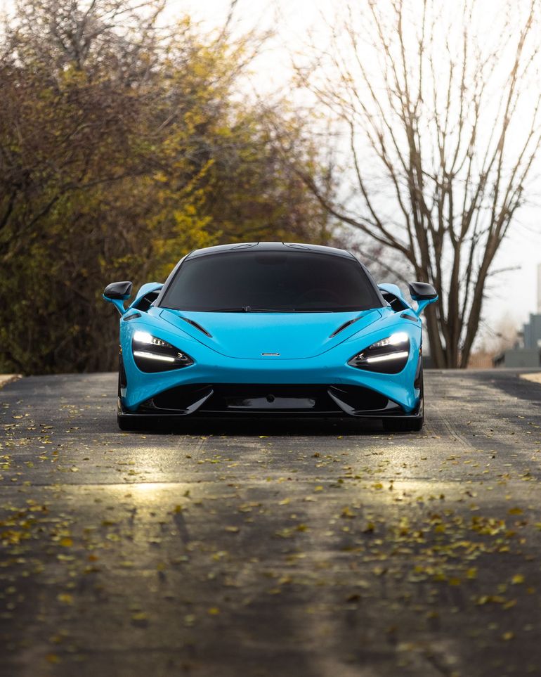 Bright blue sports car on a tree-lined road with headlights on, viewed from the front.