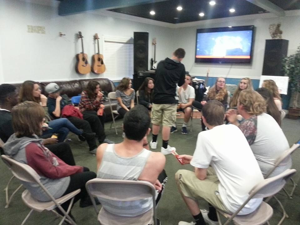 Teenagers sit in a circle, listening to a person speaking in a casual setting with guitars, speakers, and a TV.