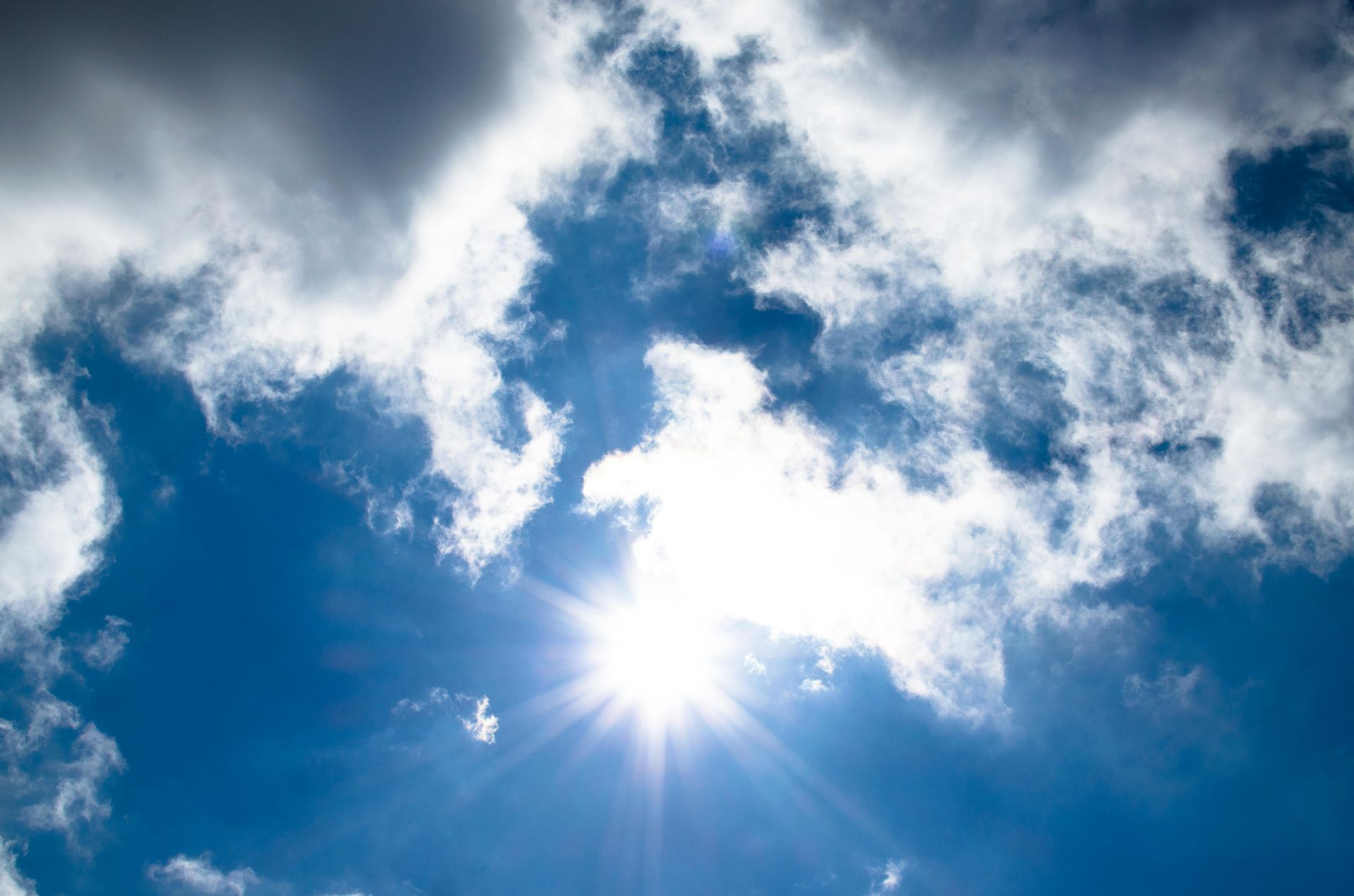 Bright sun shining through fluffy white clouds in a clear blue sky.