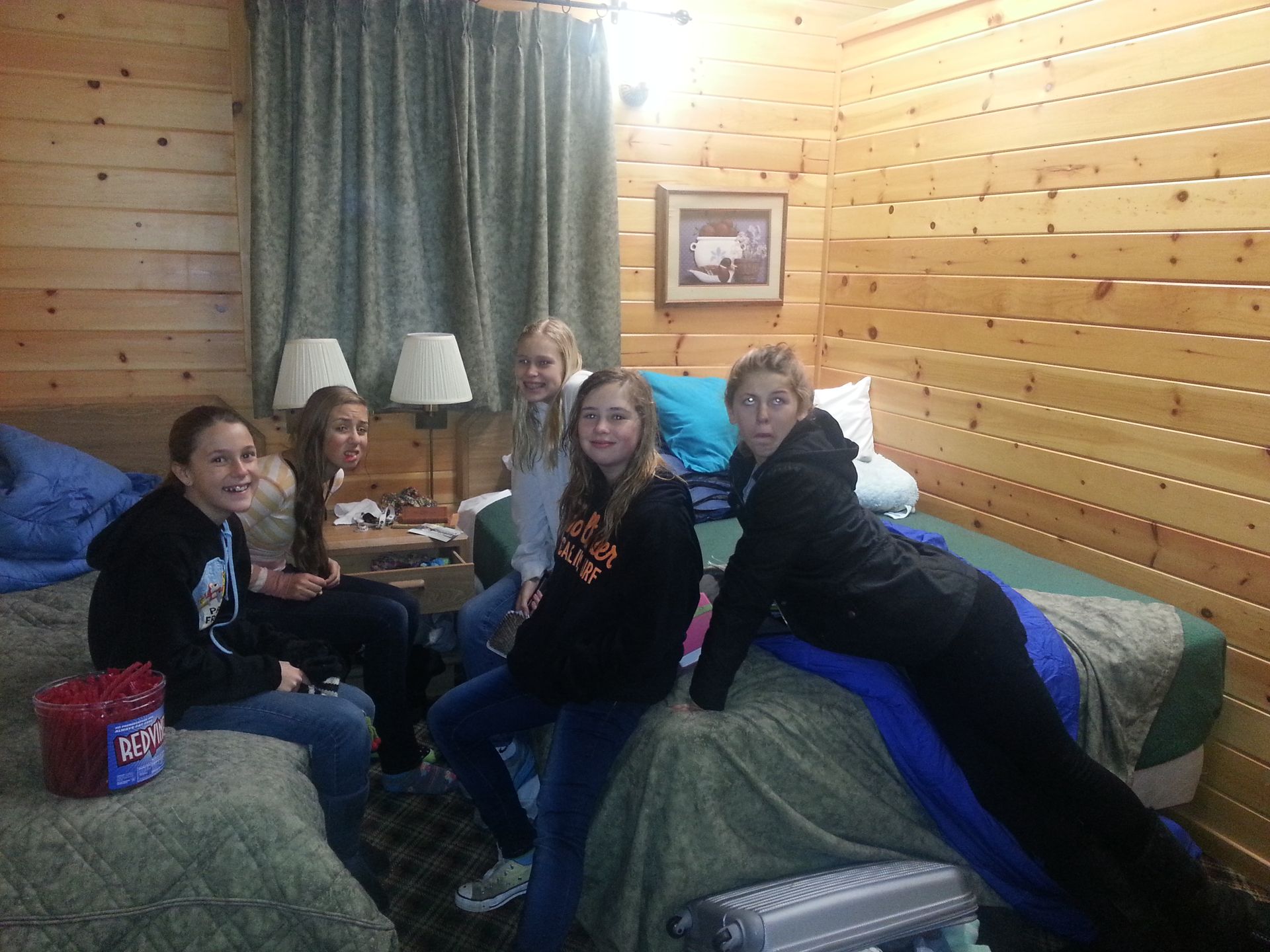 Five young people in a wood-paneled motel room, some on beds, smiling.