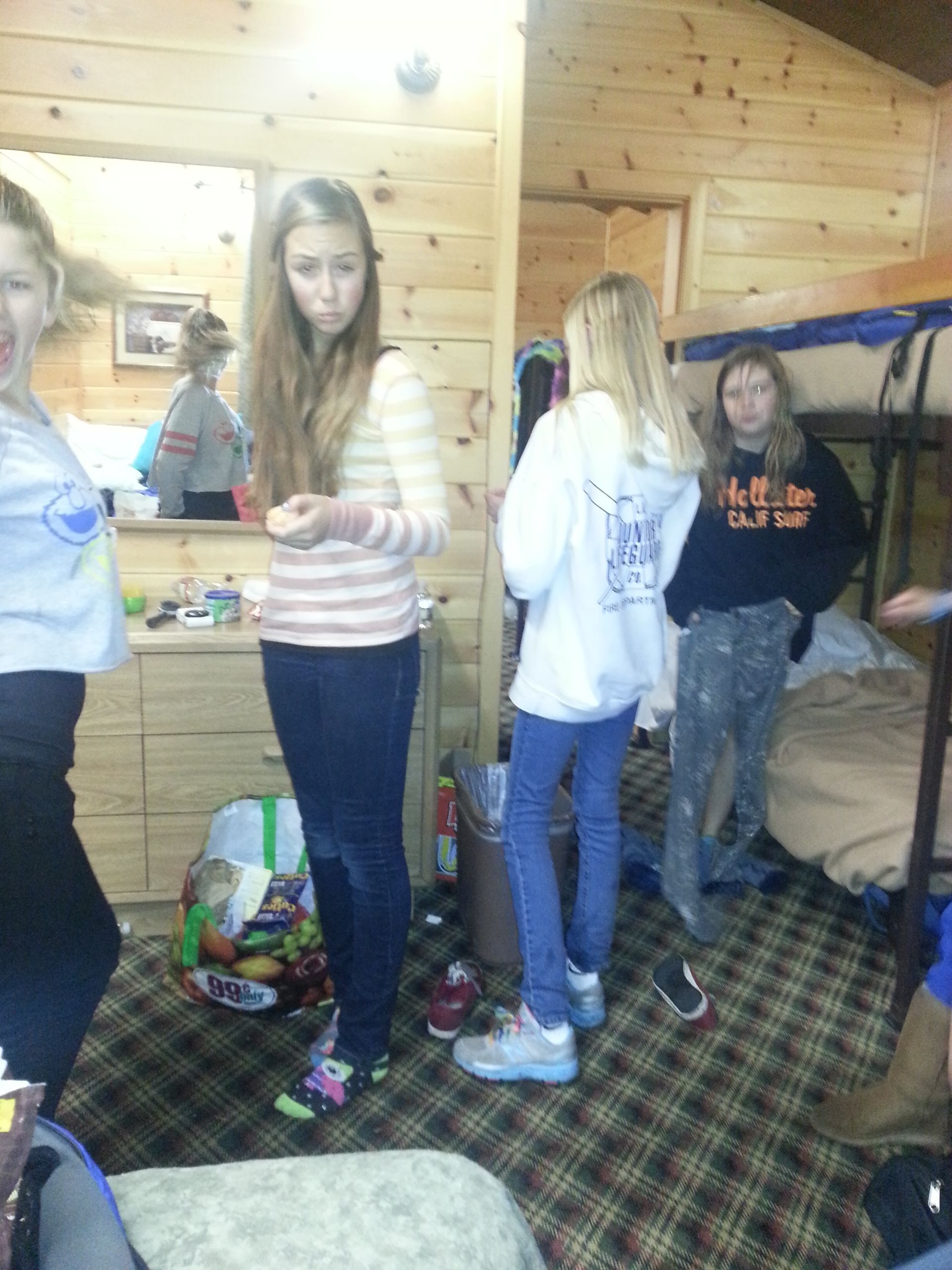 Five young women in a cabin. One in striped shirt, jeans, looks at camera. Others face away or to the side.