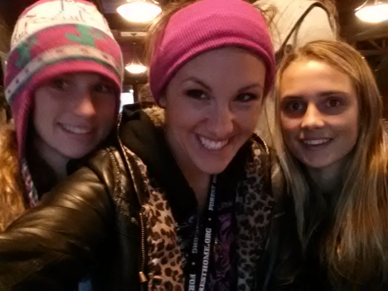 Three smiling women wearing hats and jackets pose for a selfie indoors.