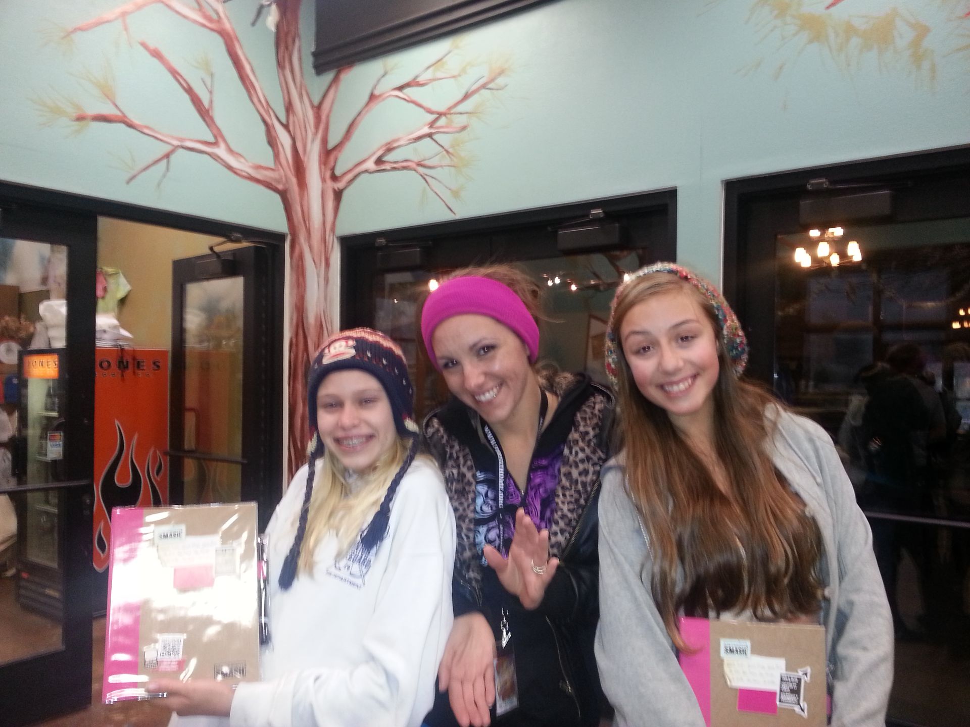Three smiling girls in hats, holding boxes. Standing in front of a door with tree mural.