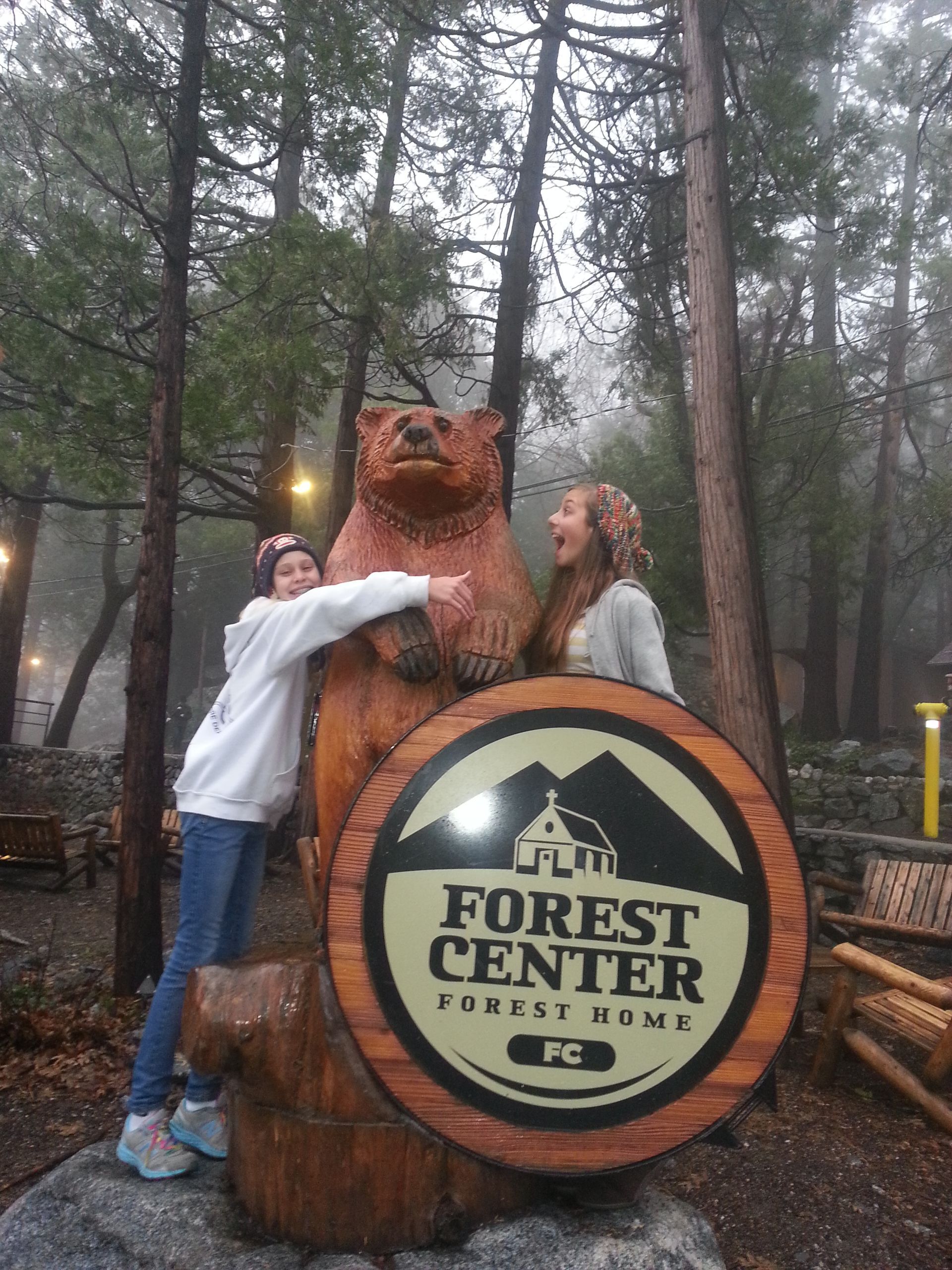 Two girls pose with a wooden bear statue next to the Forest Center sign in a forest.