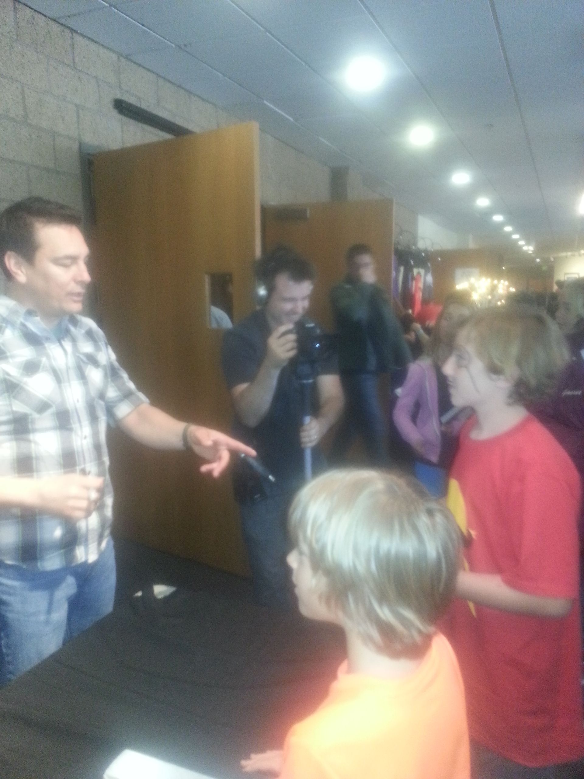 Man interviewing two boys; another man films. Interior setting, possibly a lobby.