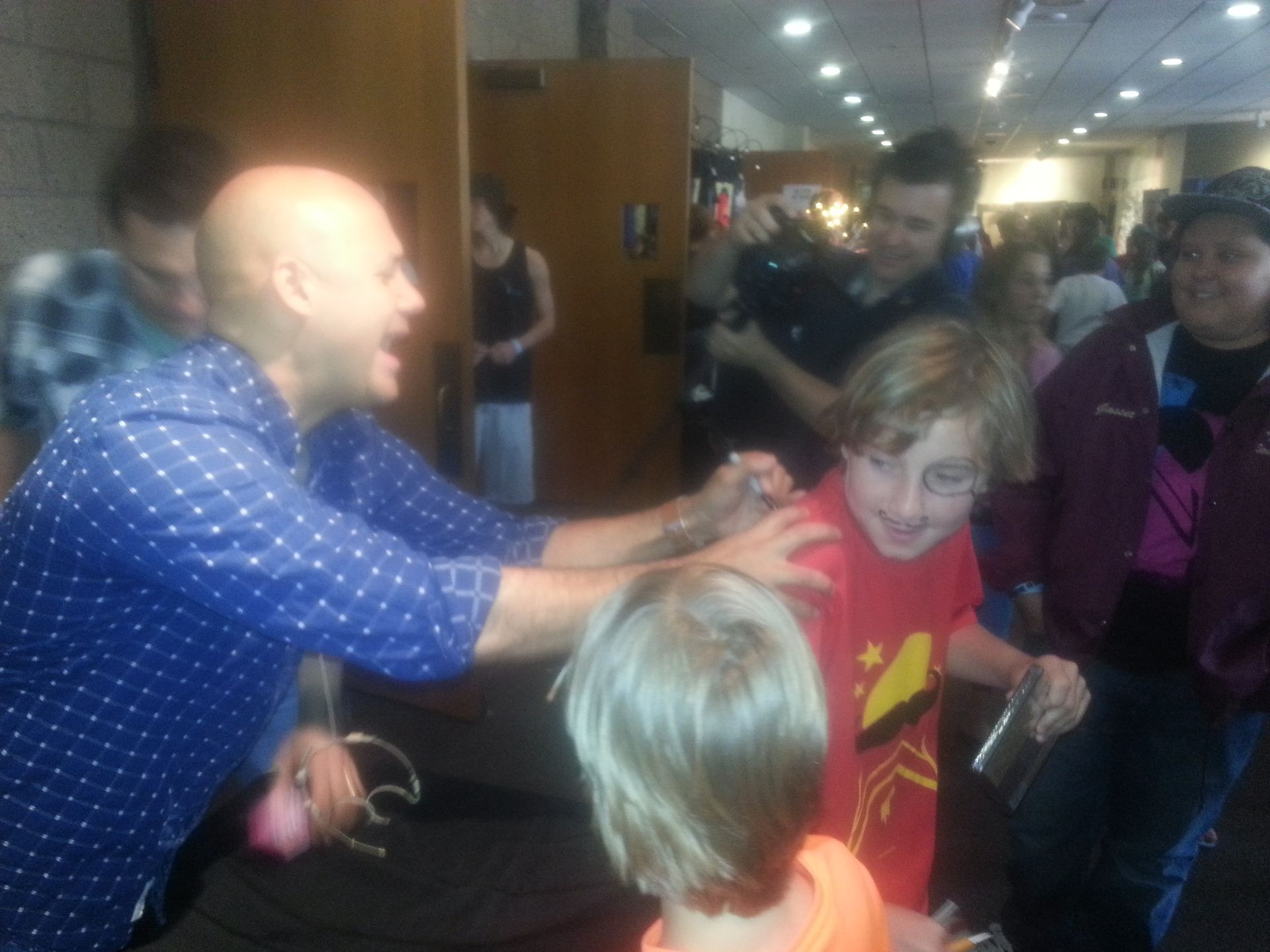 Bald man in blue shirt playfully touches a boy's shoulder in a crowded indoor space.