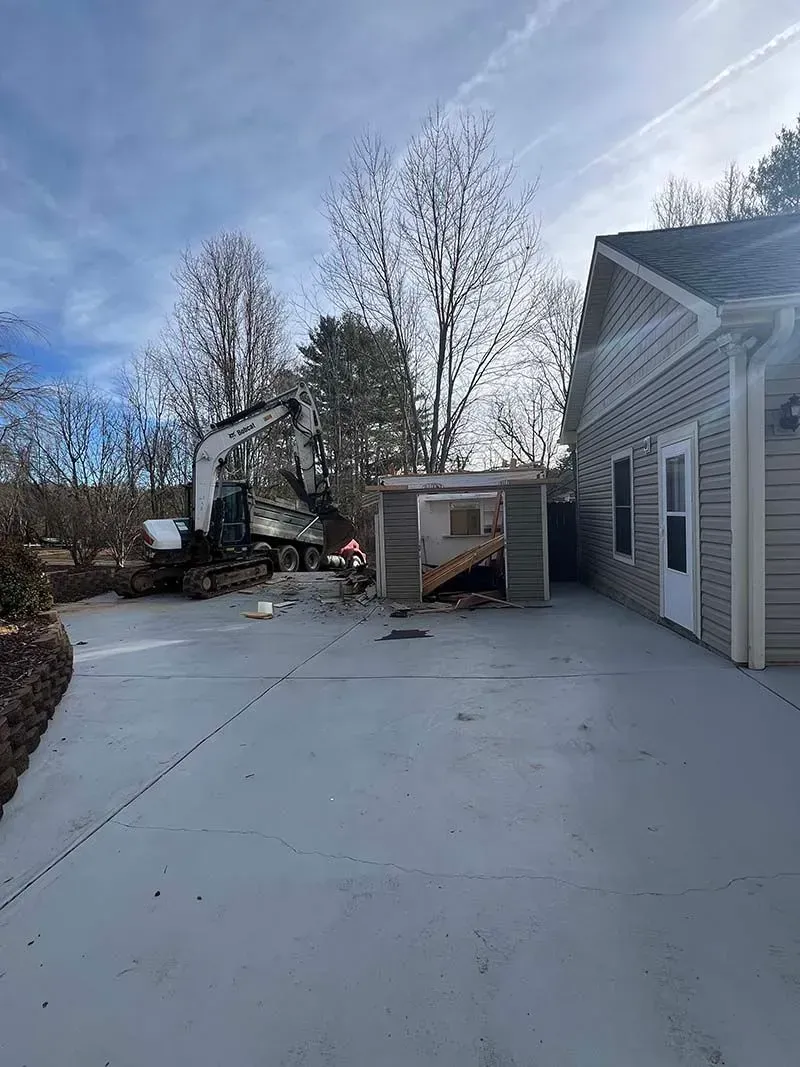 A concrete driveway leading to a house with a large excavator in the background.