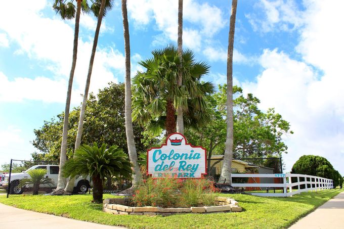 Entrance sign for Colonia del Rey resort with palm trees, lawn, and white fence under a blue sky