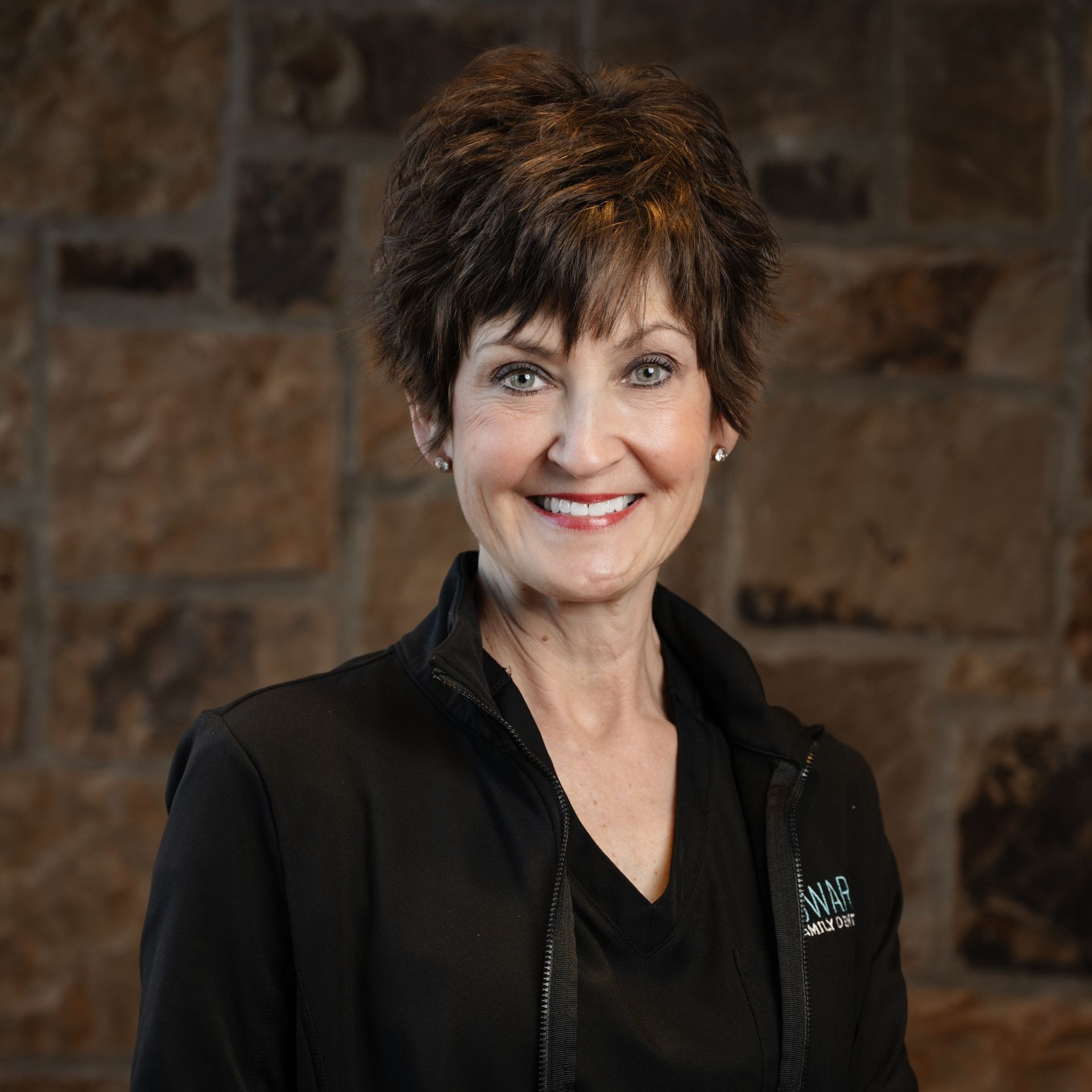 Woman with short brown hair smiles in front of a stone wall, wearing a black jacket.