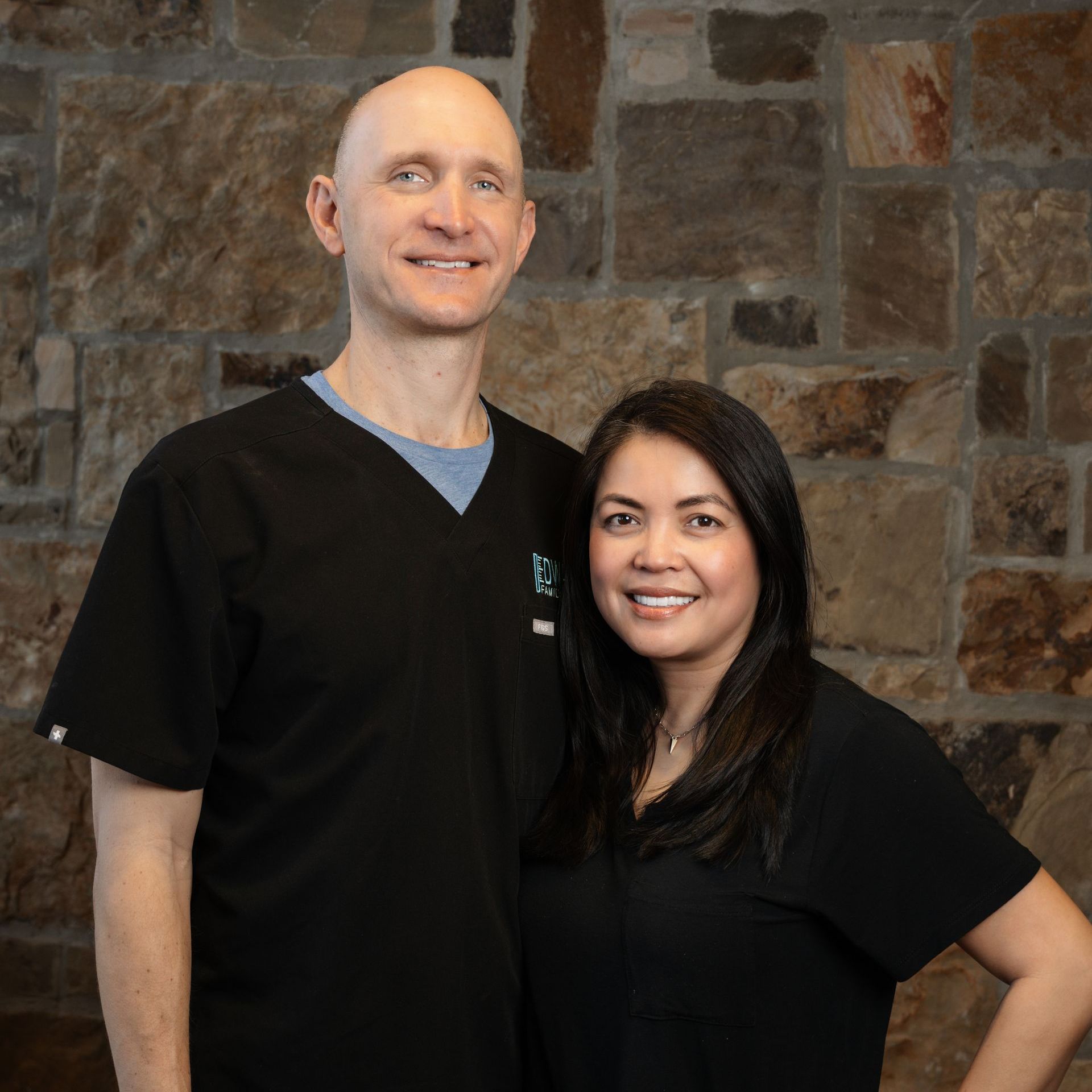 A man and a woman in black scrubs smiling in front of a stone wall.