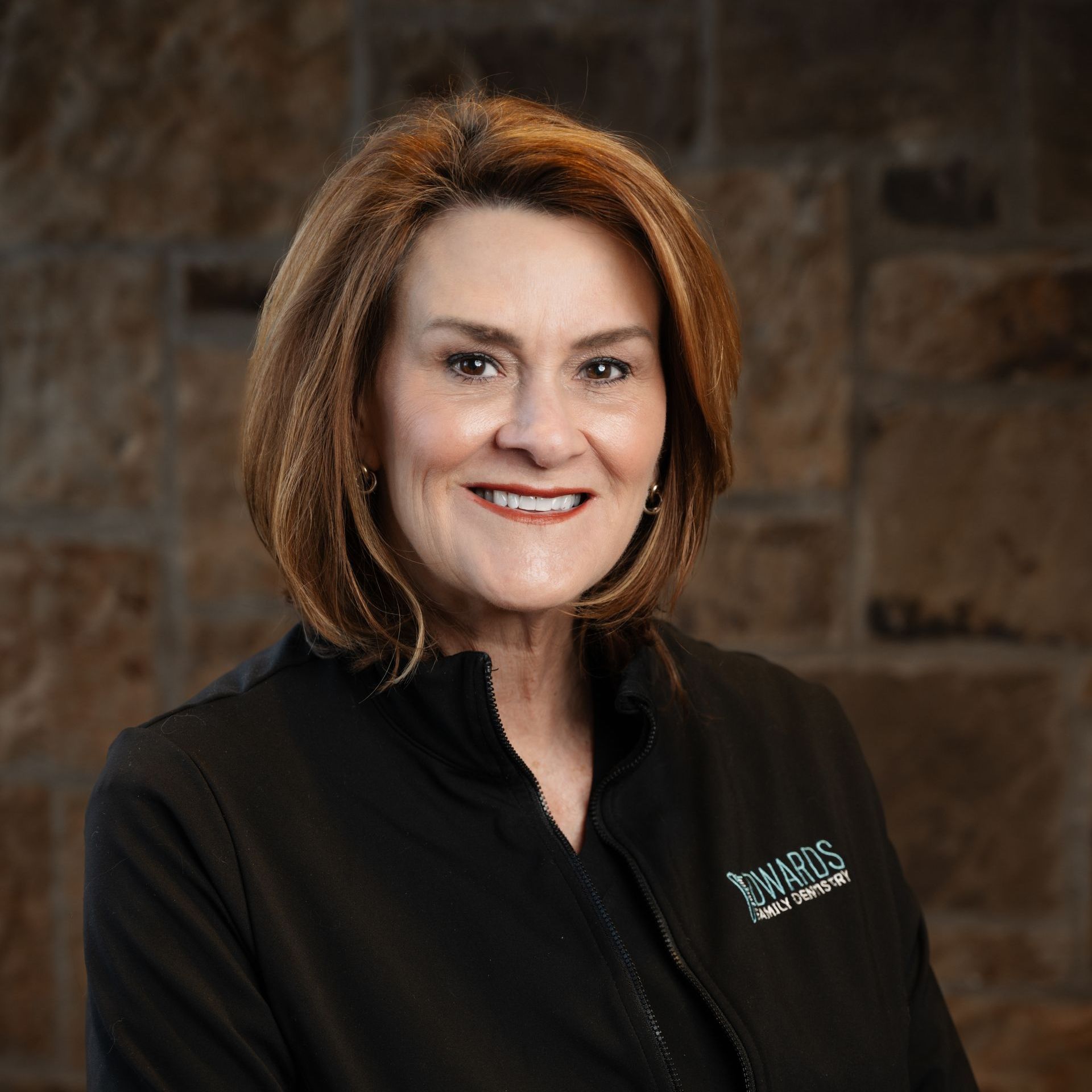 Woman in black zip-up with short brown hair smiles in front of stone wall.
