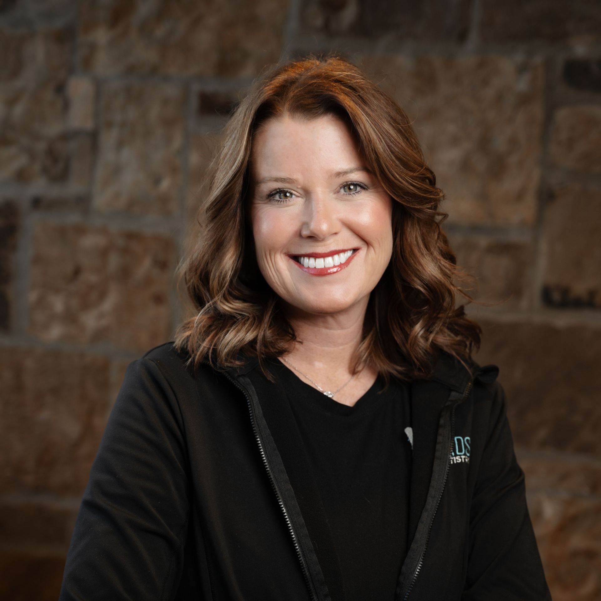 Woman with auburn hair, smiling, wearing a black jacket, in front of a stone wall.