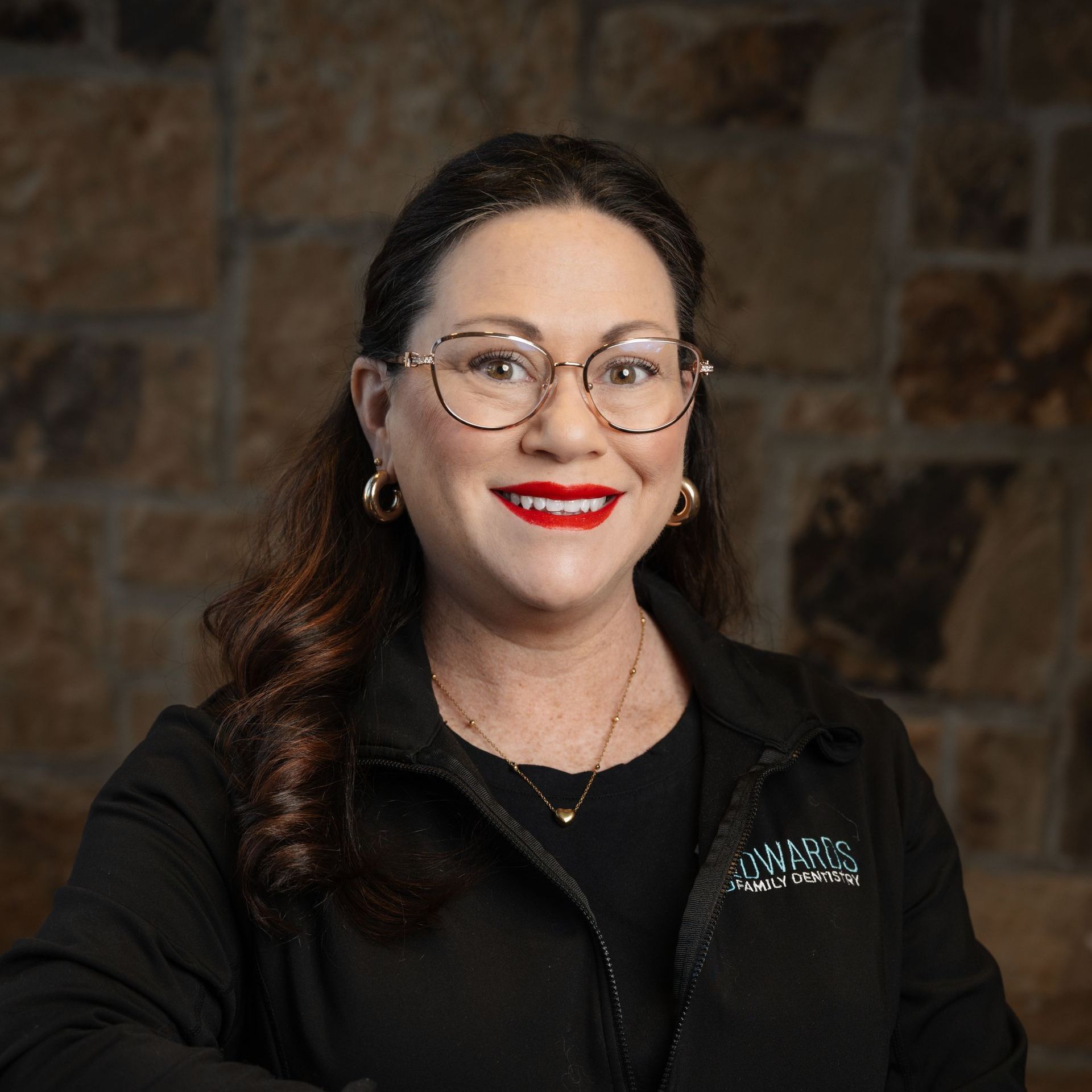 Woman with glasses and red lipstick, smiles. Wearing black jacket, gold jewelry; background is stone.