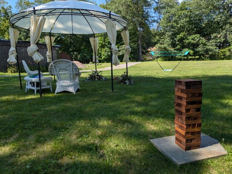 Gazebo with white curtains, wicker chairs, and a large Jenga game on a grassy lawn.