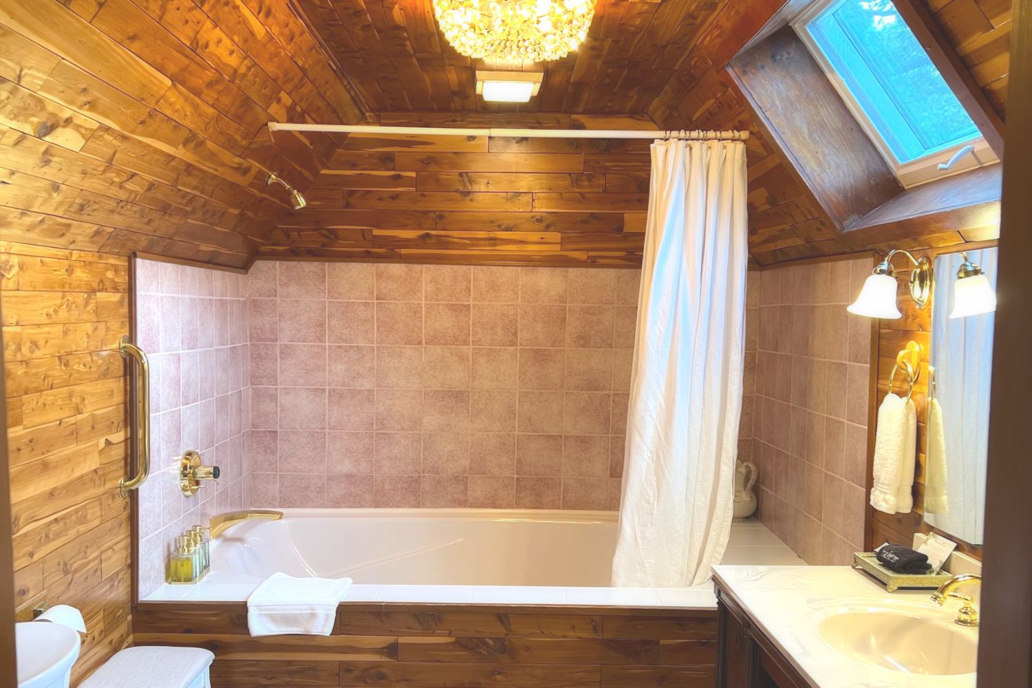 A bathroom with cedar paneling, an oversized bathtub with vintage gold hardware, a sink, mirror, and skylight.