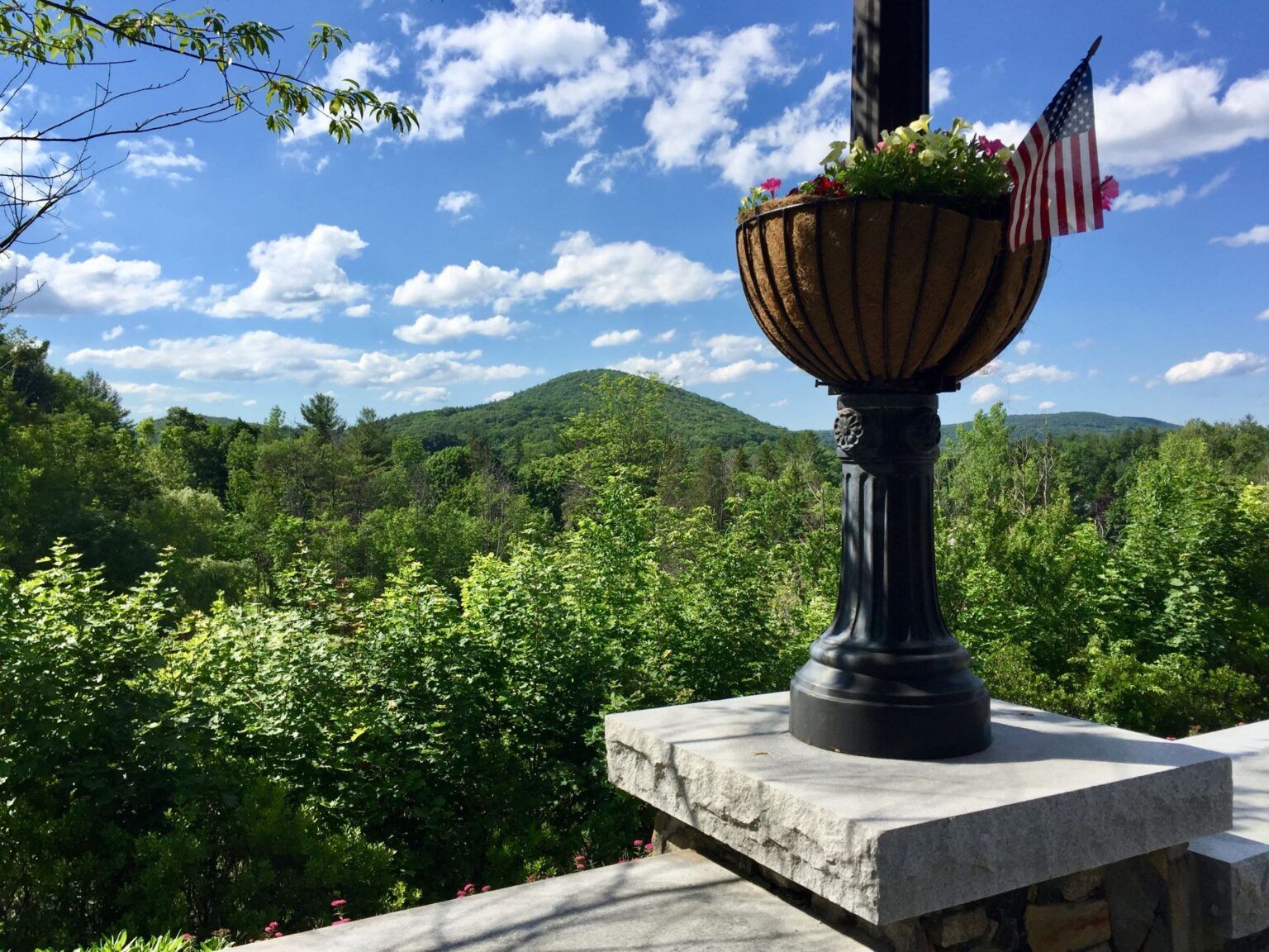 A planter with flowers and an american flag is sitting on top of a stone ledge overlooking a forest.
